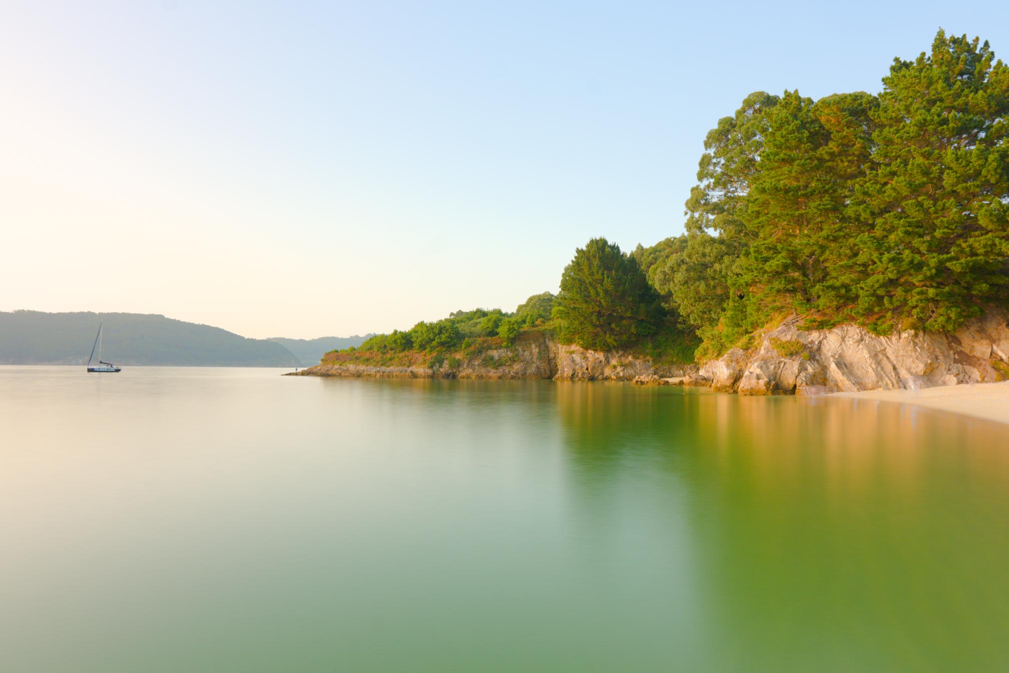 Long exposure seascape of Vidreiro Beach in Galicia, Spain, featuring calm emerald waters, a sailboat, and lush coastal cliffs.